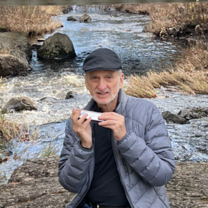 Author Richard Andrews sitting on a rock, in the middle of a stream, holding a mouth harp, with a bridge behind him